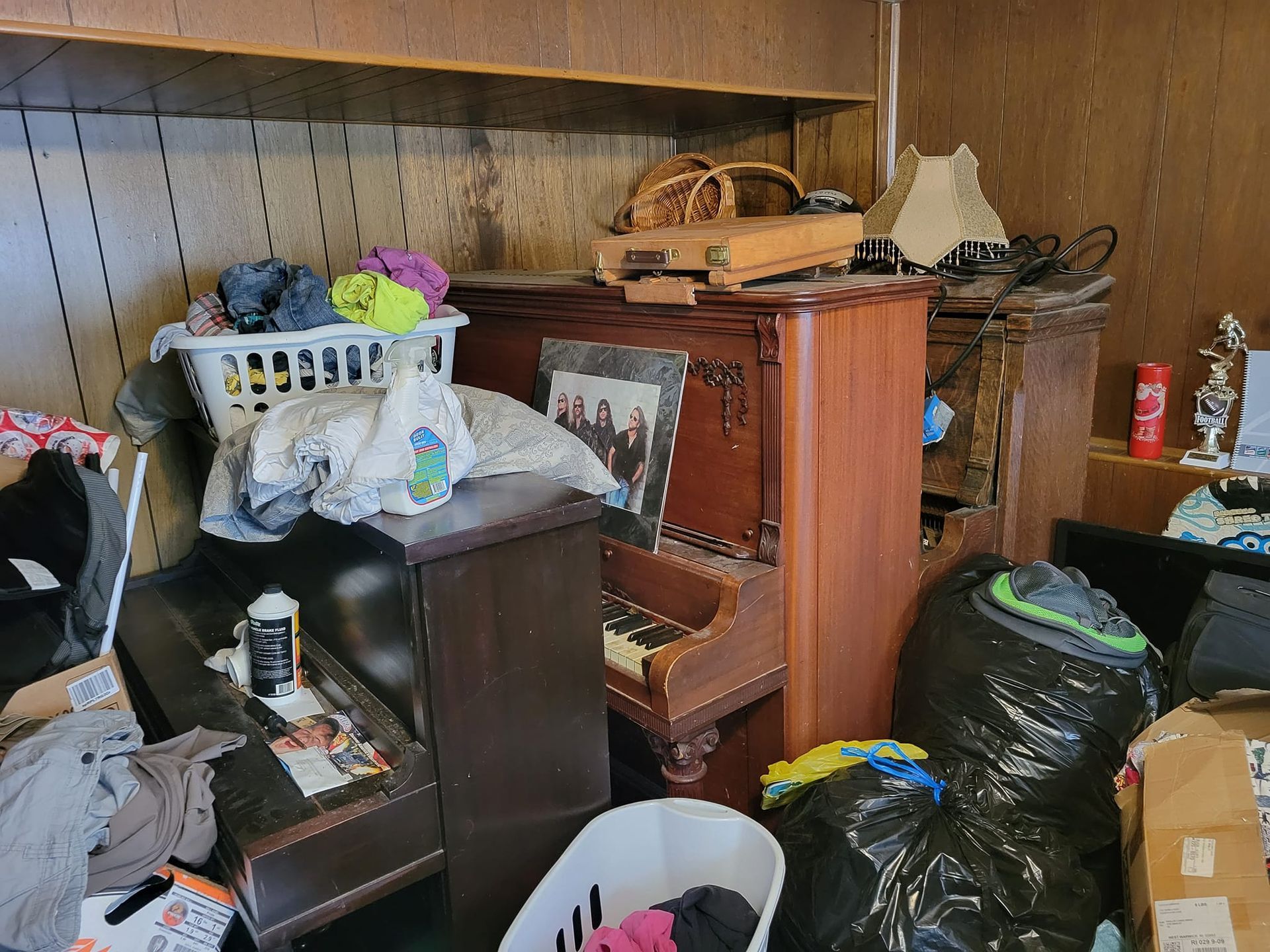 Cluttered room with a piano, laundry baskets, garbage bags, and various items; interior.