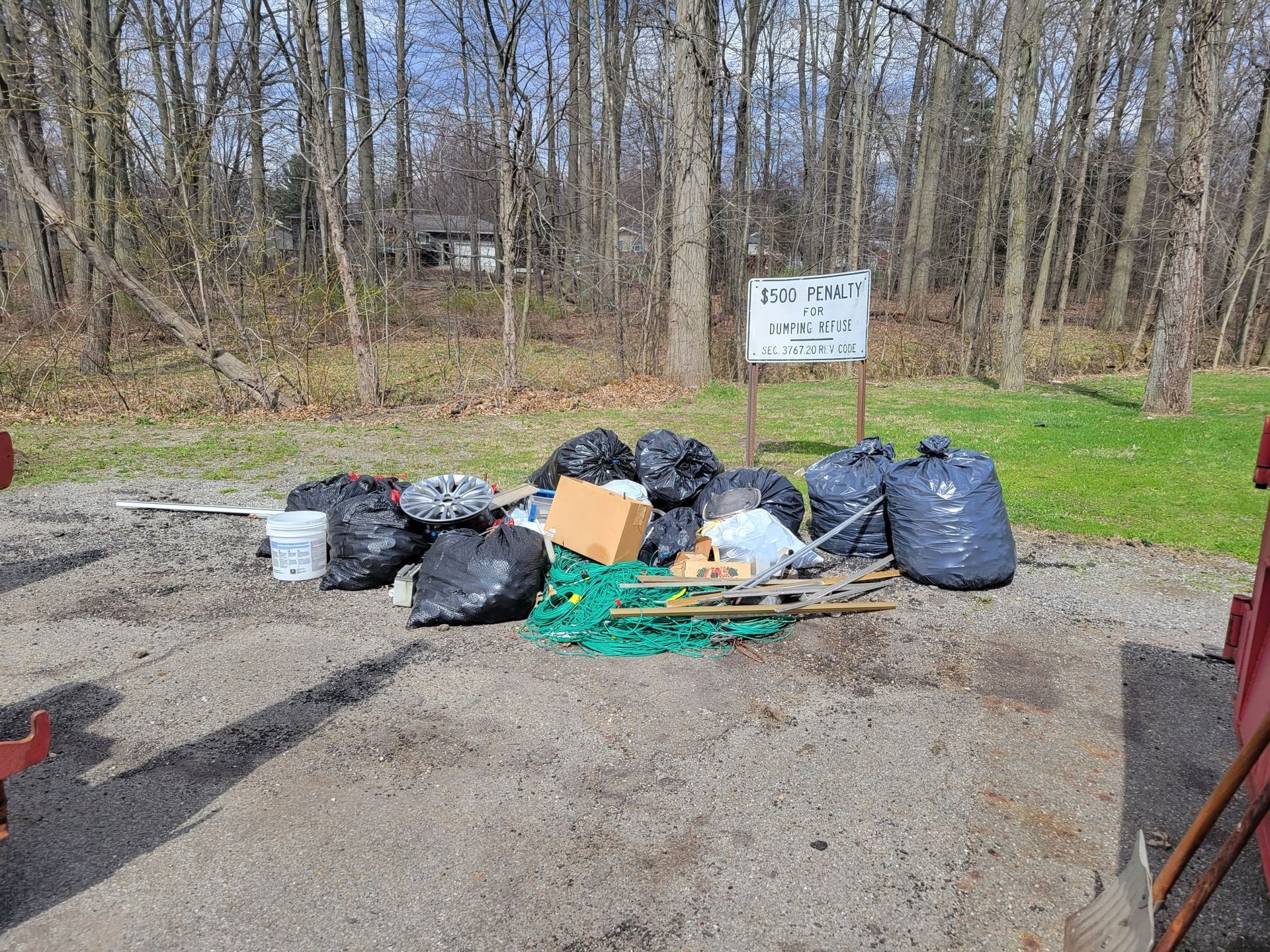 Pile of black trash bags, cardboard, and debris on gravel with a sign in a wooded area.