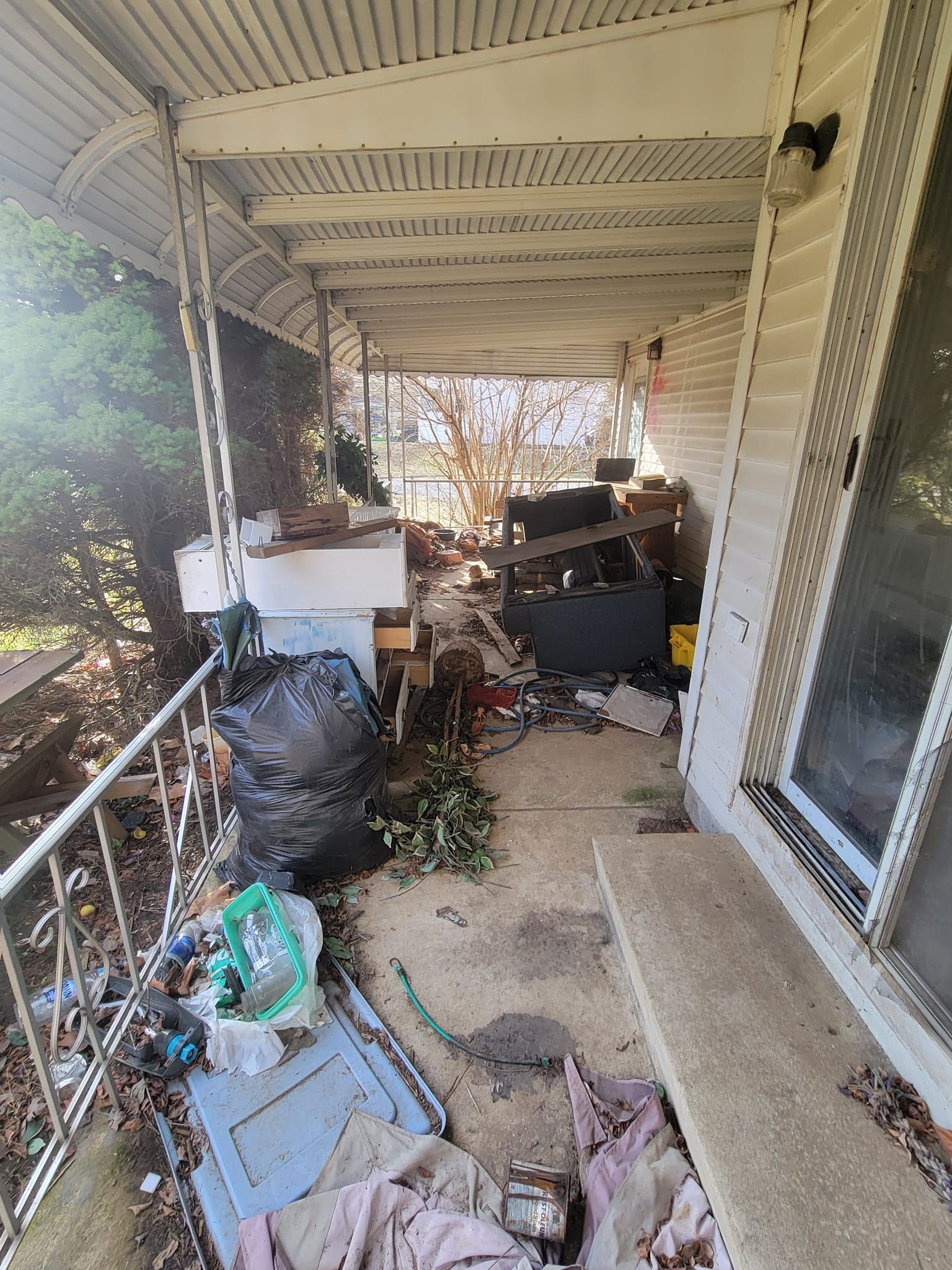 Overgrown porch cluttered with debris, trash bags, and overgrown foliage.