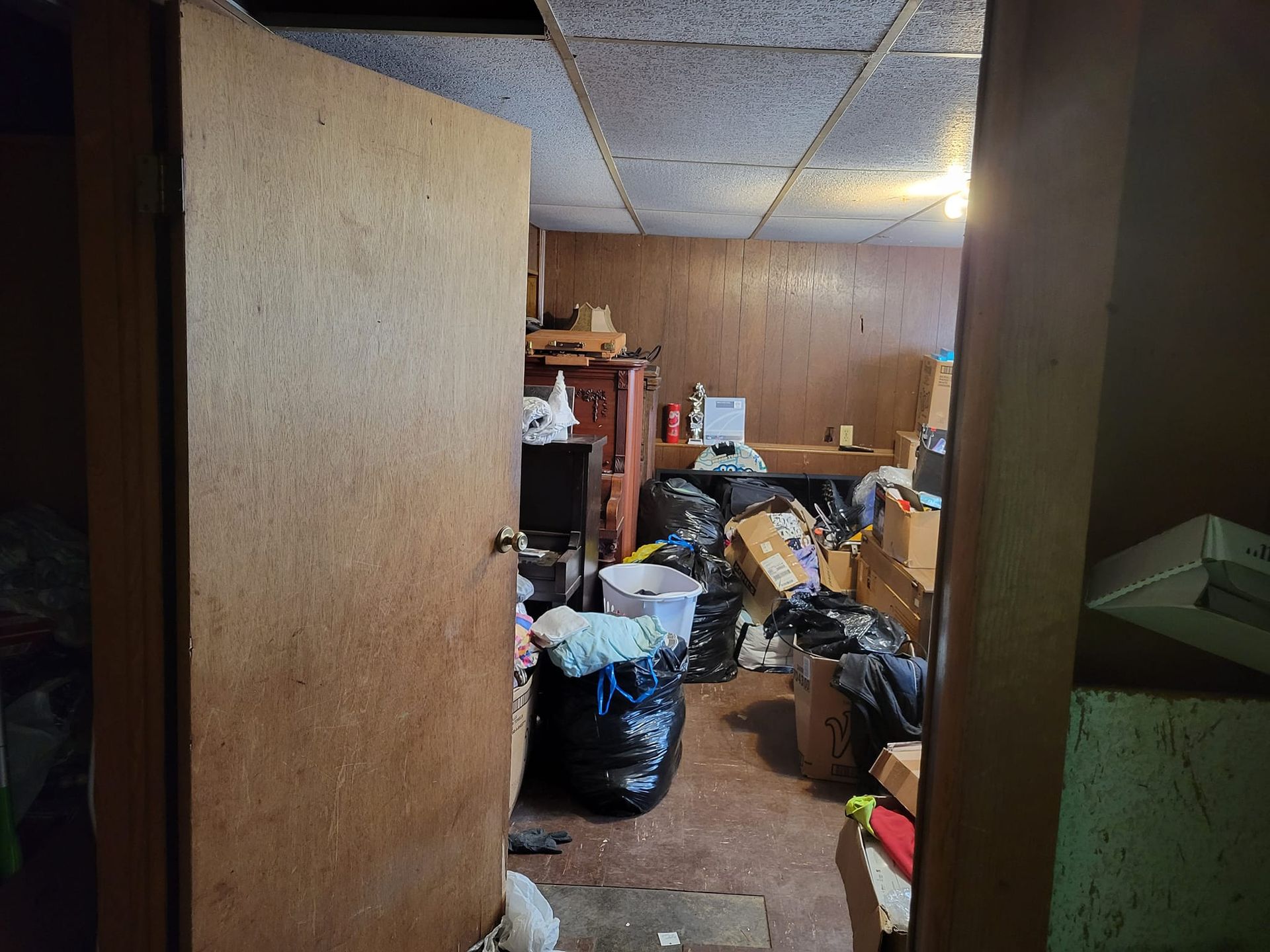 Doorway into a cluttered room: trash bags, boxes, furniture, and a drop ceiling.