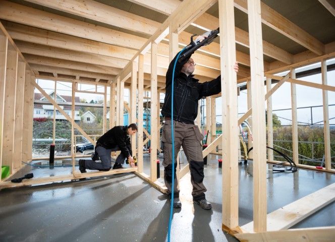 Two people building a wooden frame structure indoors. One uses a nail gun. Light wood, gray floor.