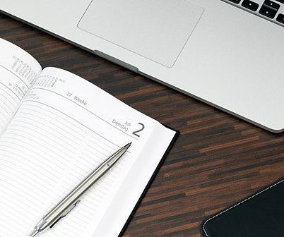 A pen is sitting on top of an open notebook next to a laptop.