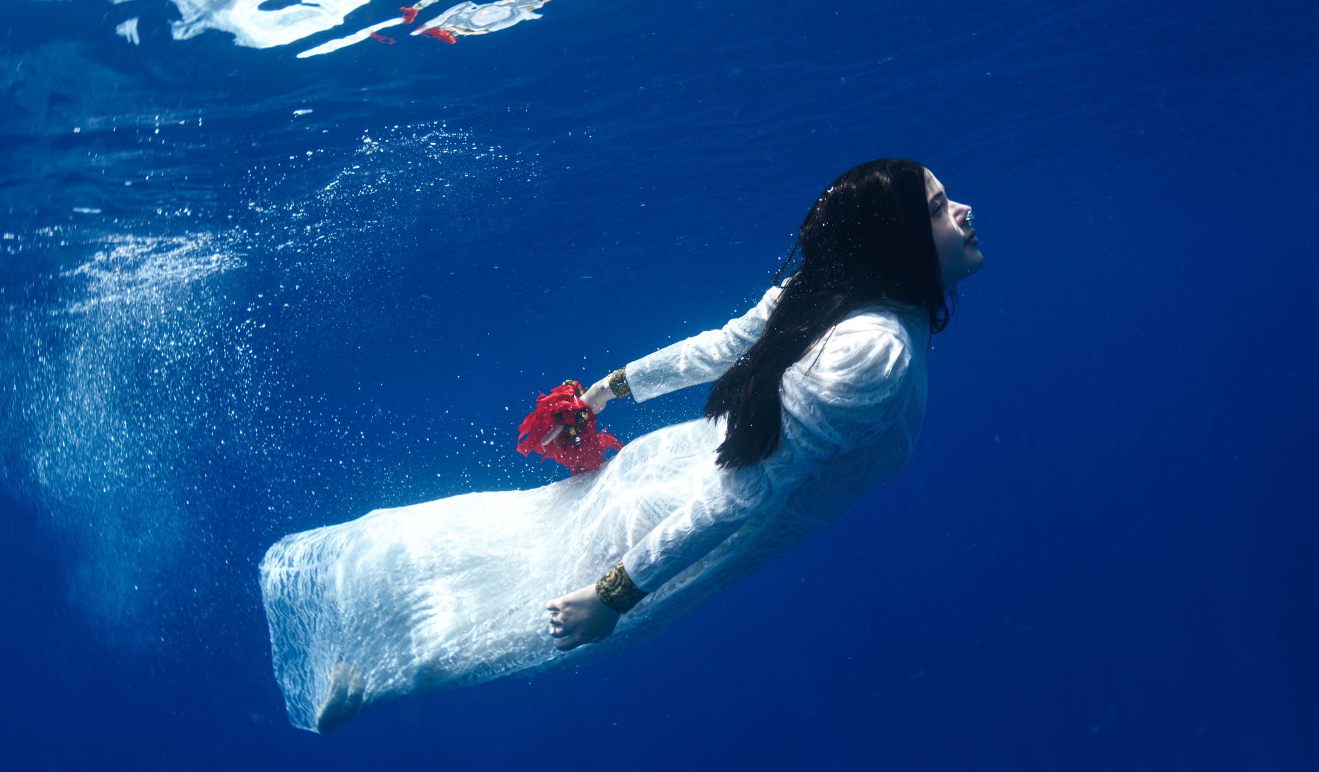 A Fiji bride wearing a long white wedding dress and holding a red flower swims through darker blue water