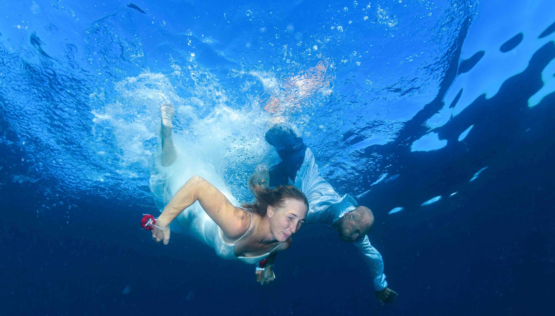 A Fiji wedding couple  holding hands dive into clear blue water creating a wave of bubbles and swimming downwards