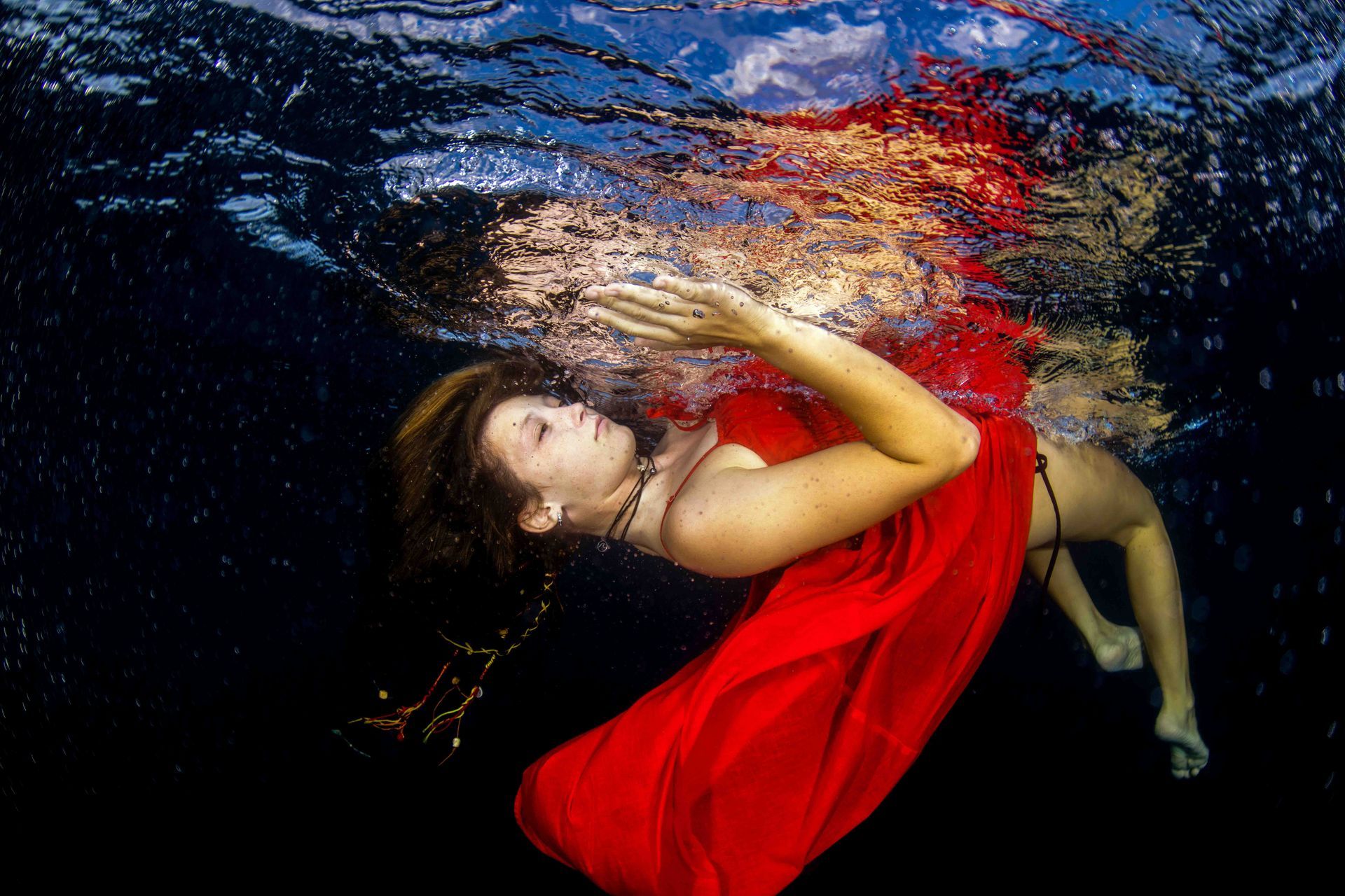 A fine art portrait of a young girl wearing a red dress lying under the surface of the water and creatIng a stunning reflection in Fiji 