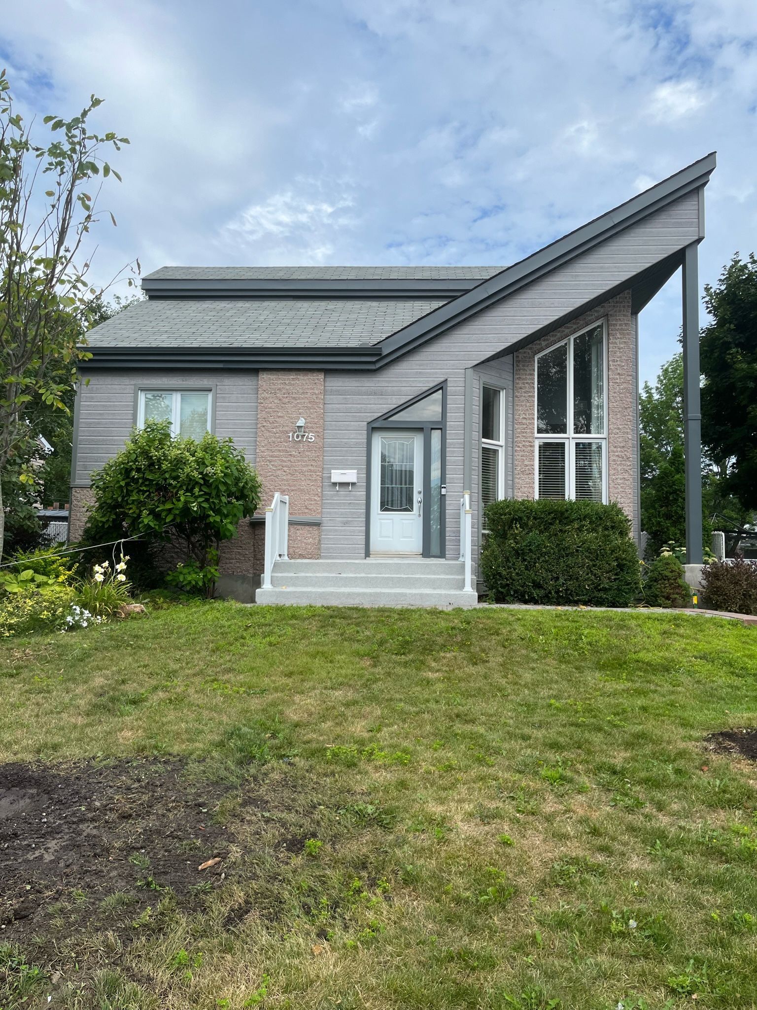 Maison moderne avec toit gris en pente, éléments en brique, porte blanche et jardin gazonné.