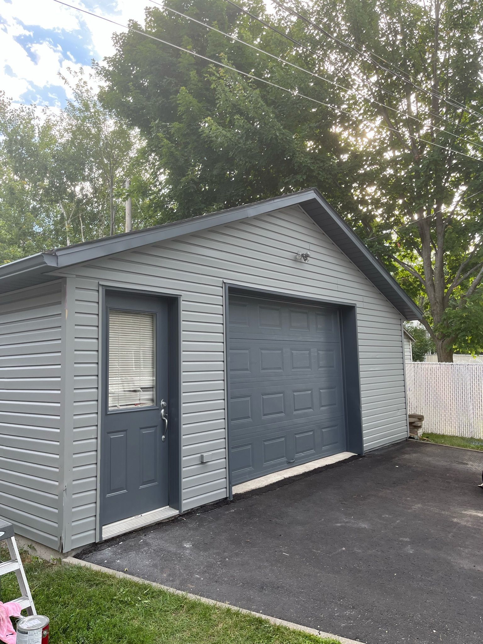 Garage gris avec porte et porte de garage gris foncé, allée asphaltée et clôture blanche.