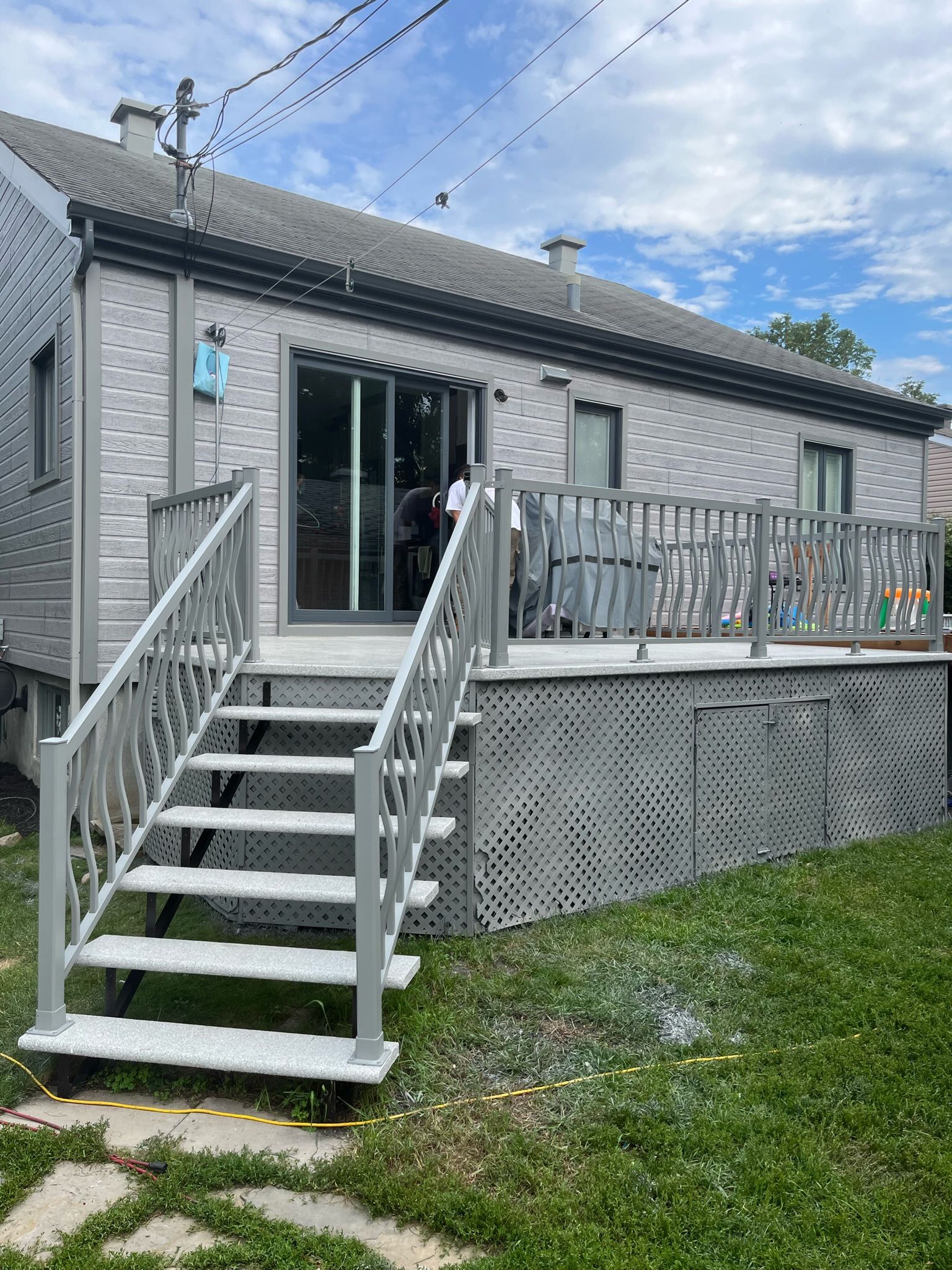 Une terrasse grise et un escalier mènent à une maison aux portes coulissantes en verre. Ciel couvert.