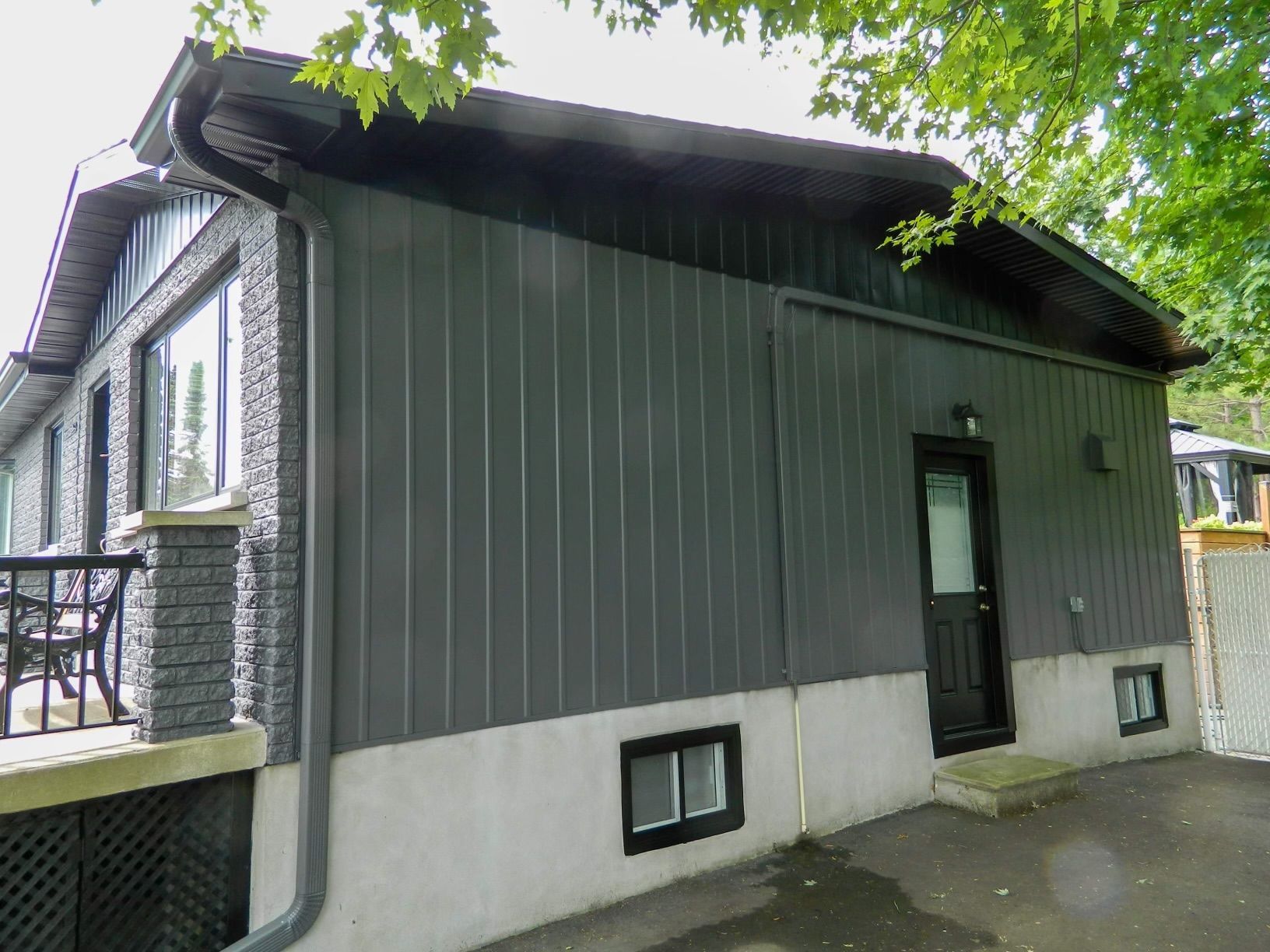 Maison gris foncé avec porte noire, fondations en béton et petites fenêtres.