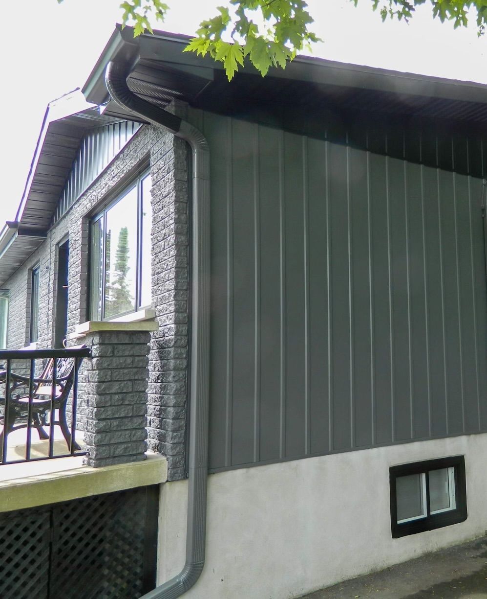 Maison à l'extérieur gris et en pierre, avec lambris vertical, gouttière et petite fenêtre au sous-sol.