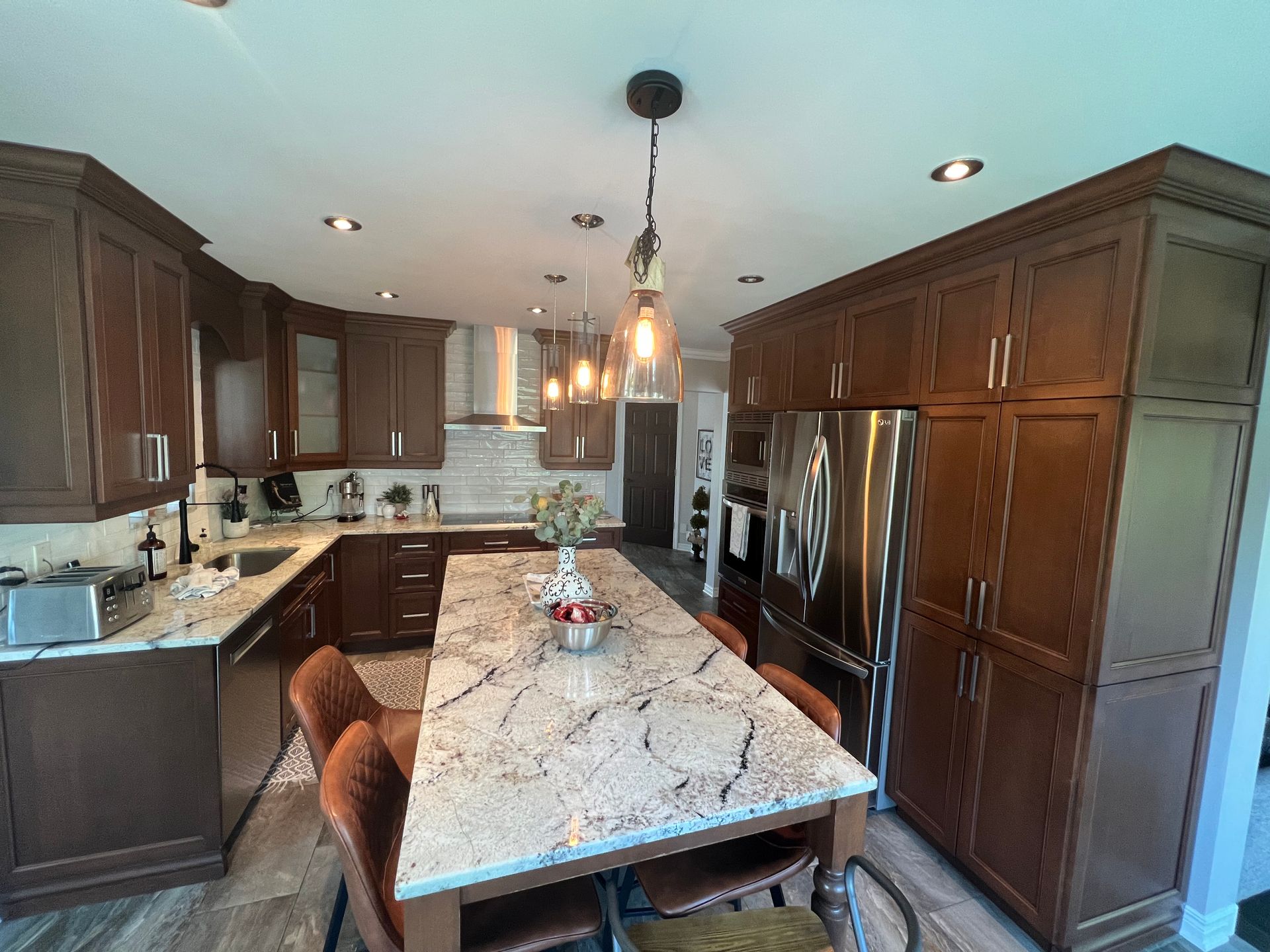 Vue grand angle d'une cuisine moderne avec des armoires en bois foncé, un grand îlot en granit, des appareils électroménagers en acier inoxydable et des suspensions lumineuses.