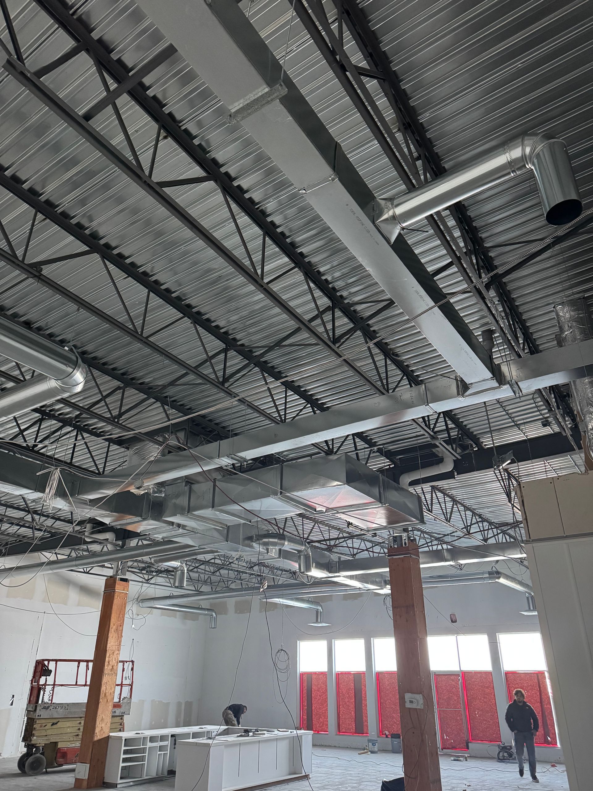 Scène de chantier montrant des conduits métalliques suspendus à un plafond apparent, avec des ouvriers et du matériel dans la pièce.