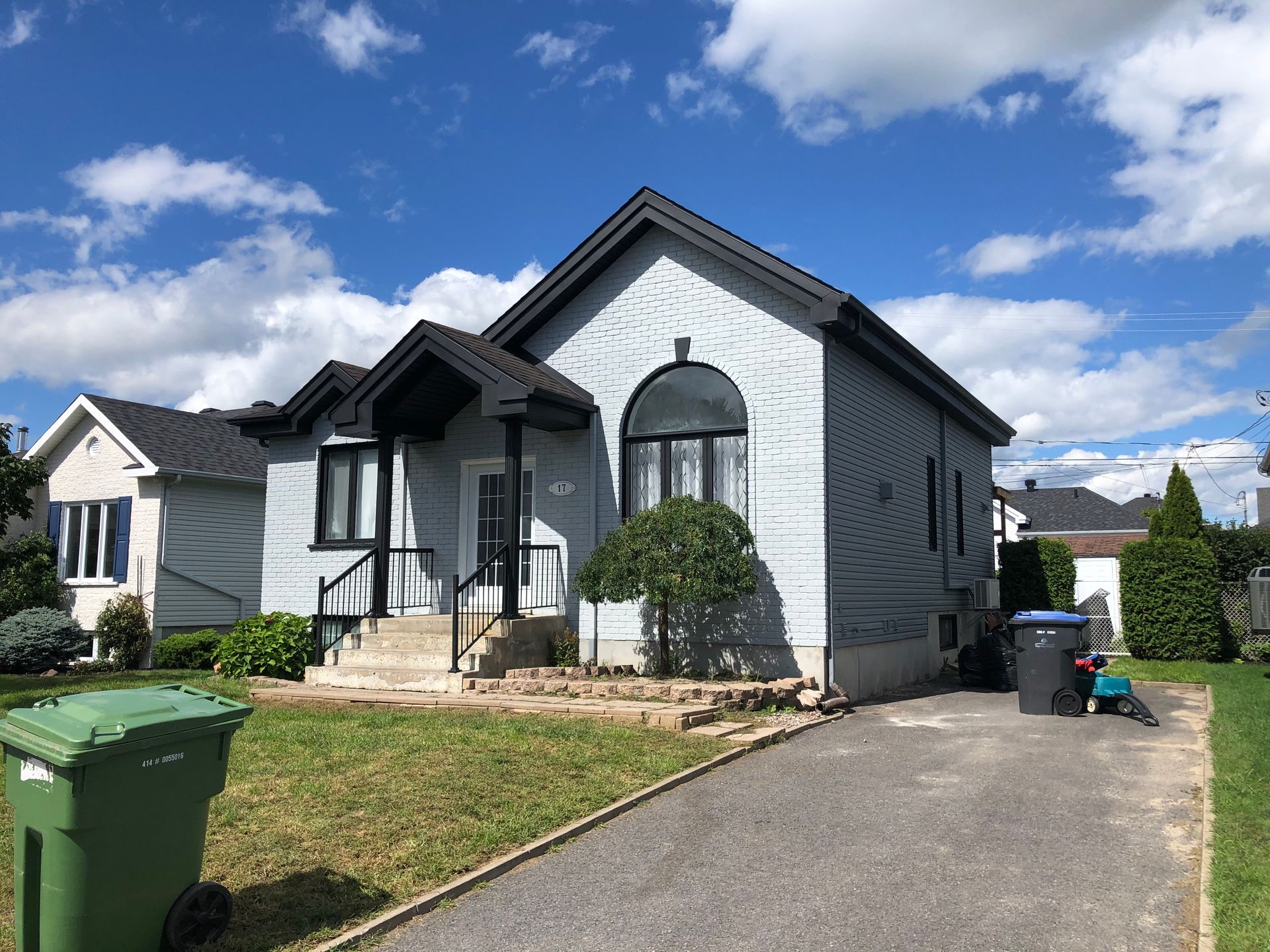 une maison blanche avec un toit noir et une poubelle verte devant.