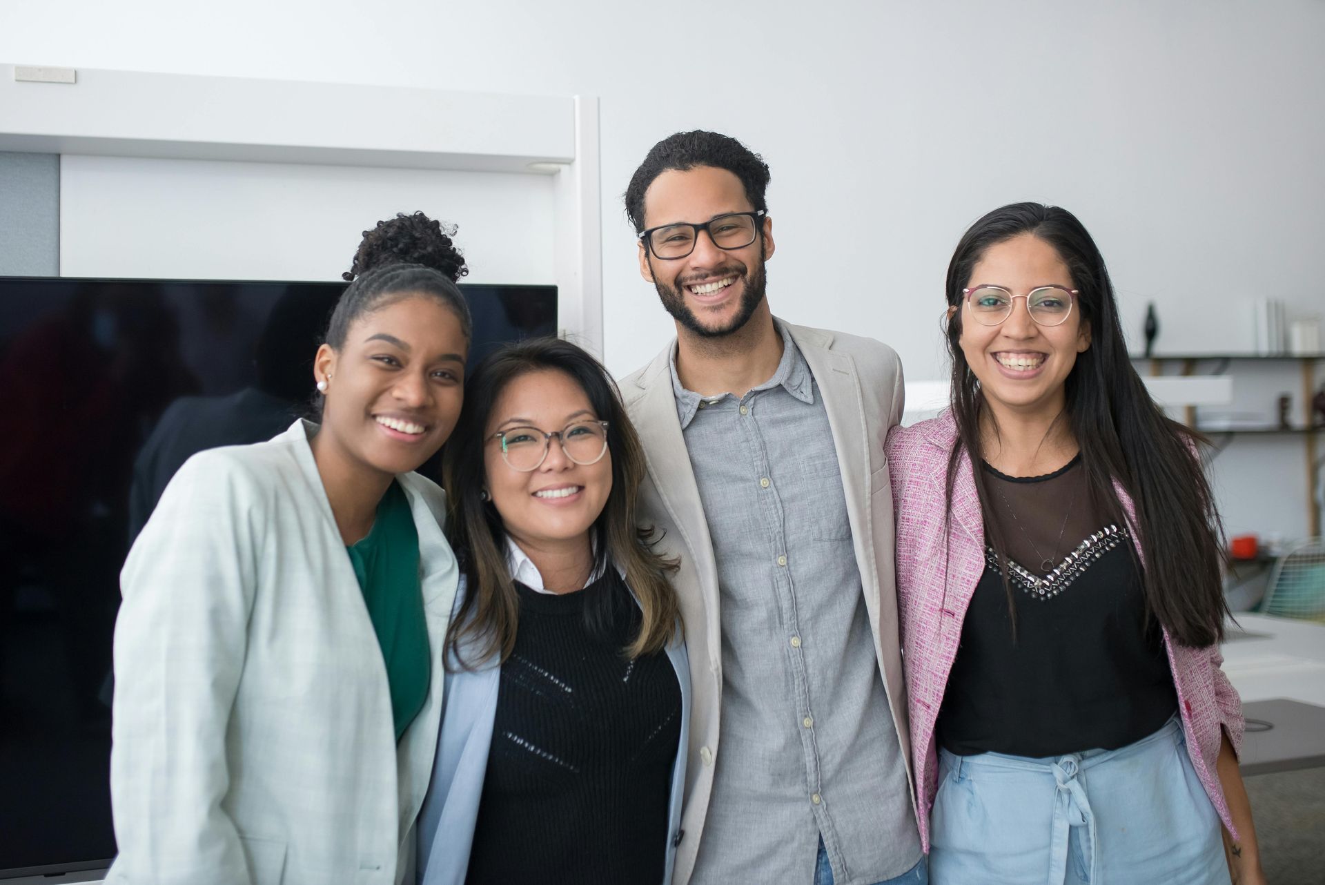 Four smiling people posing together in an office setting; a man in a blazer, and three women.
