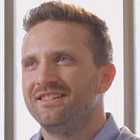 Woman with brown hair smiles broadly outdoors, greenery in background.