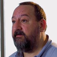Man with a beard, looking slightly left. Wearing a blue collared shirt, against a bright background.