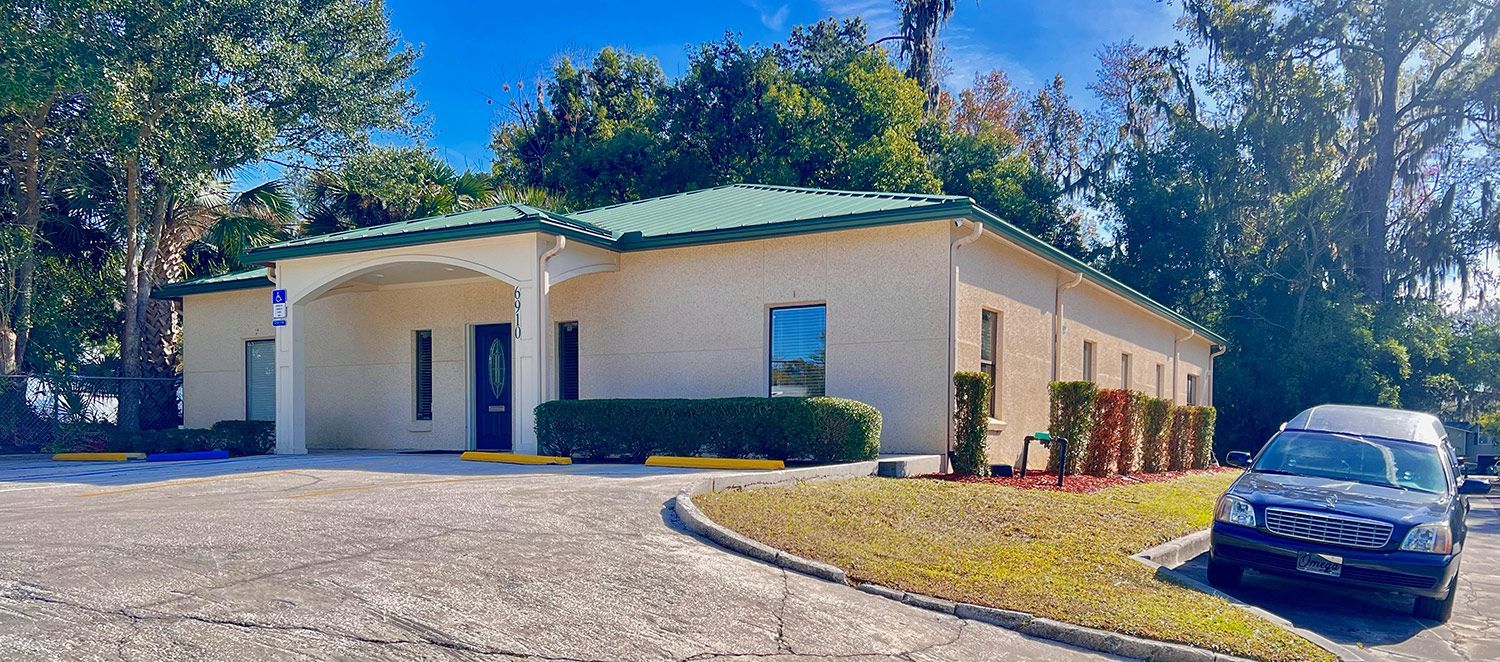 A one-story beige building with a dark green roof, a covered entrance, and a gravel parking lot with a parked car.