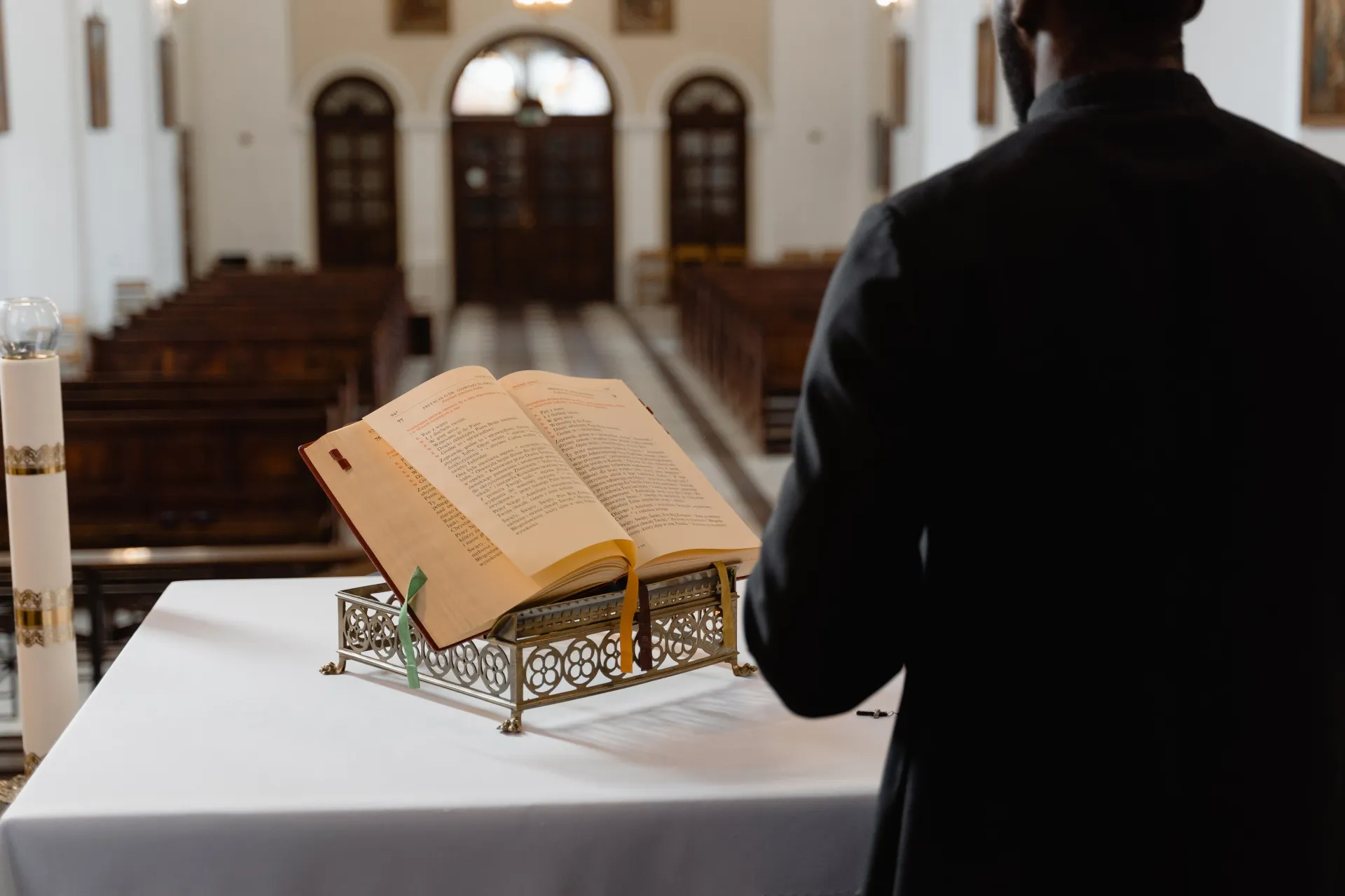 A person in a suit stands at a lectern in a church, looking toward an open book on a decorative stand.