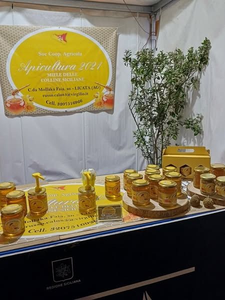 Vasetti di miele esposti in una mostra di Apicultura 2021, con un cartello e decorazioni vegetali.