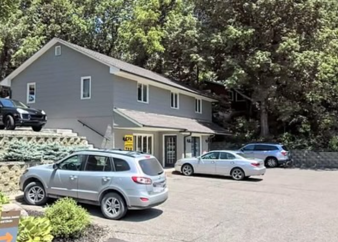 A two-story gray building with several parked cars in a parking lot on a sunny day.