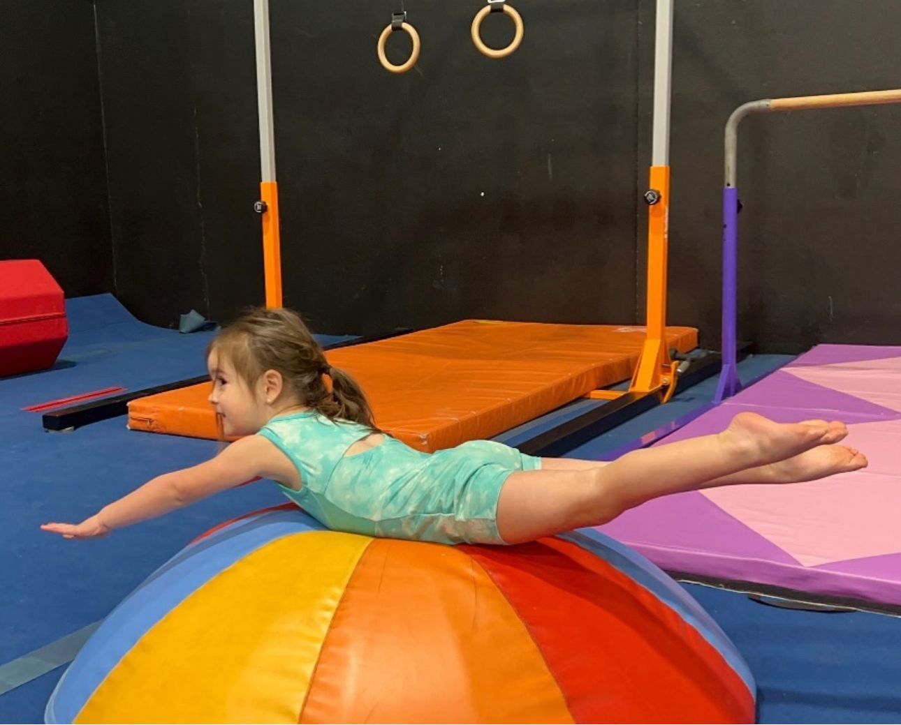 A little girl is laying on a colorful ball in a gym.