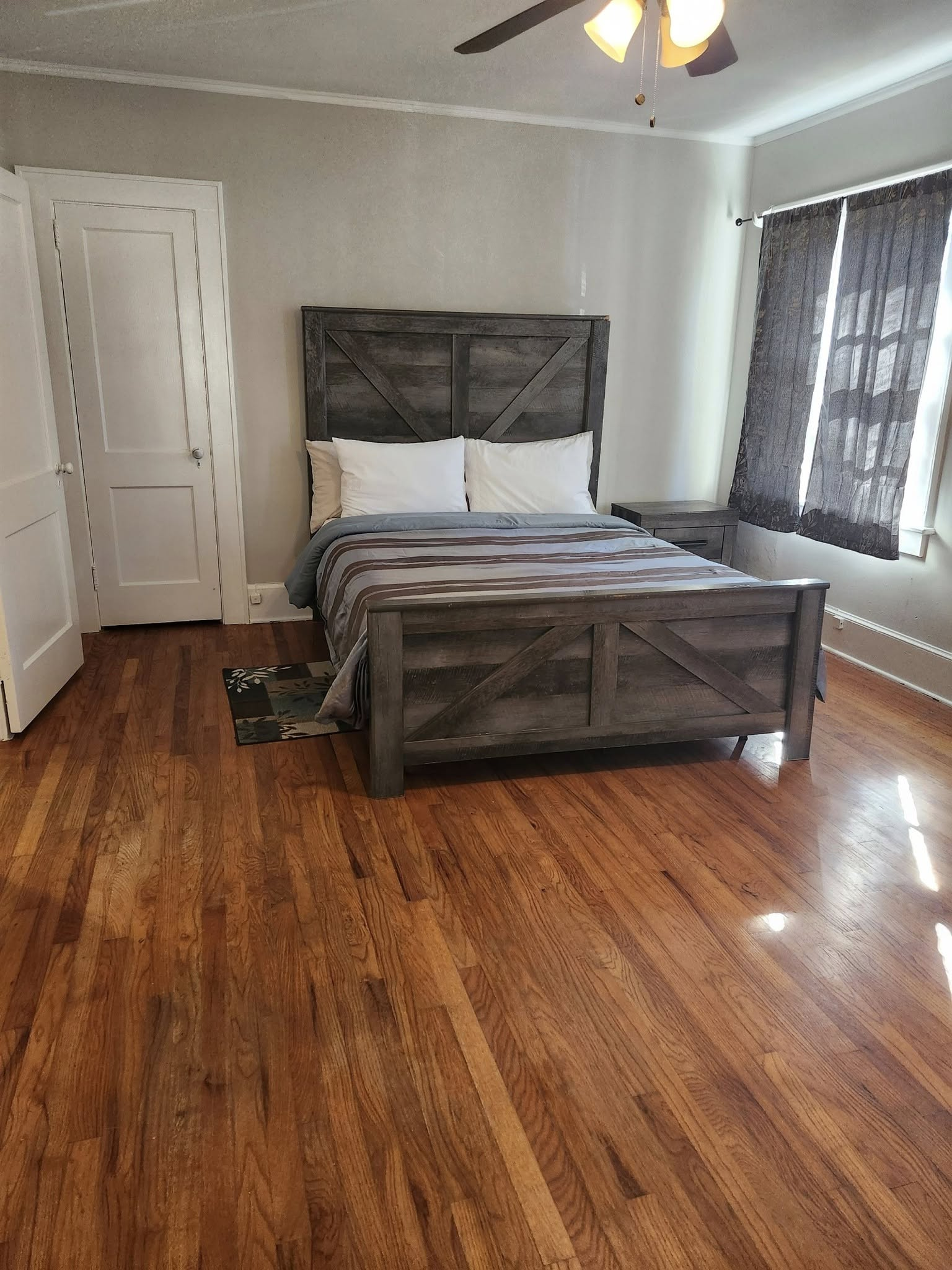 Bedroom with wooden floor, grey bed, nightstand, and window with curtains.