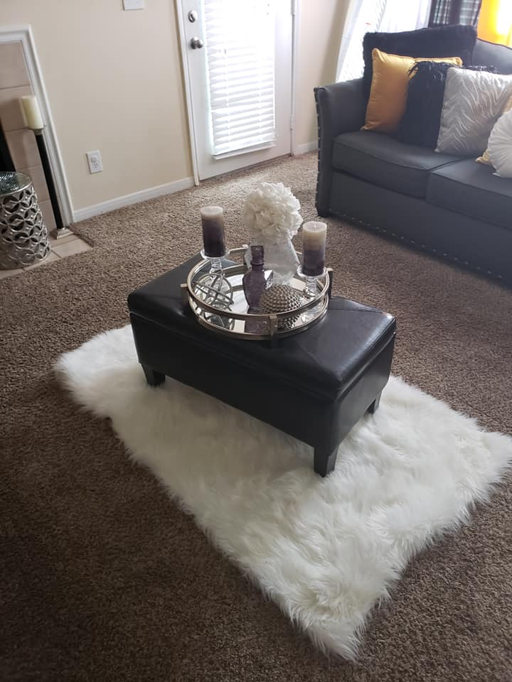 Black ottoman with tray, candles, and flowers on a white faux fur rug on brown carpet in a living room.