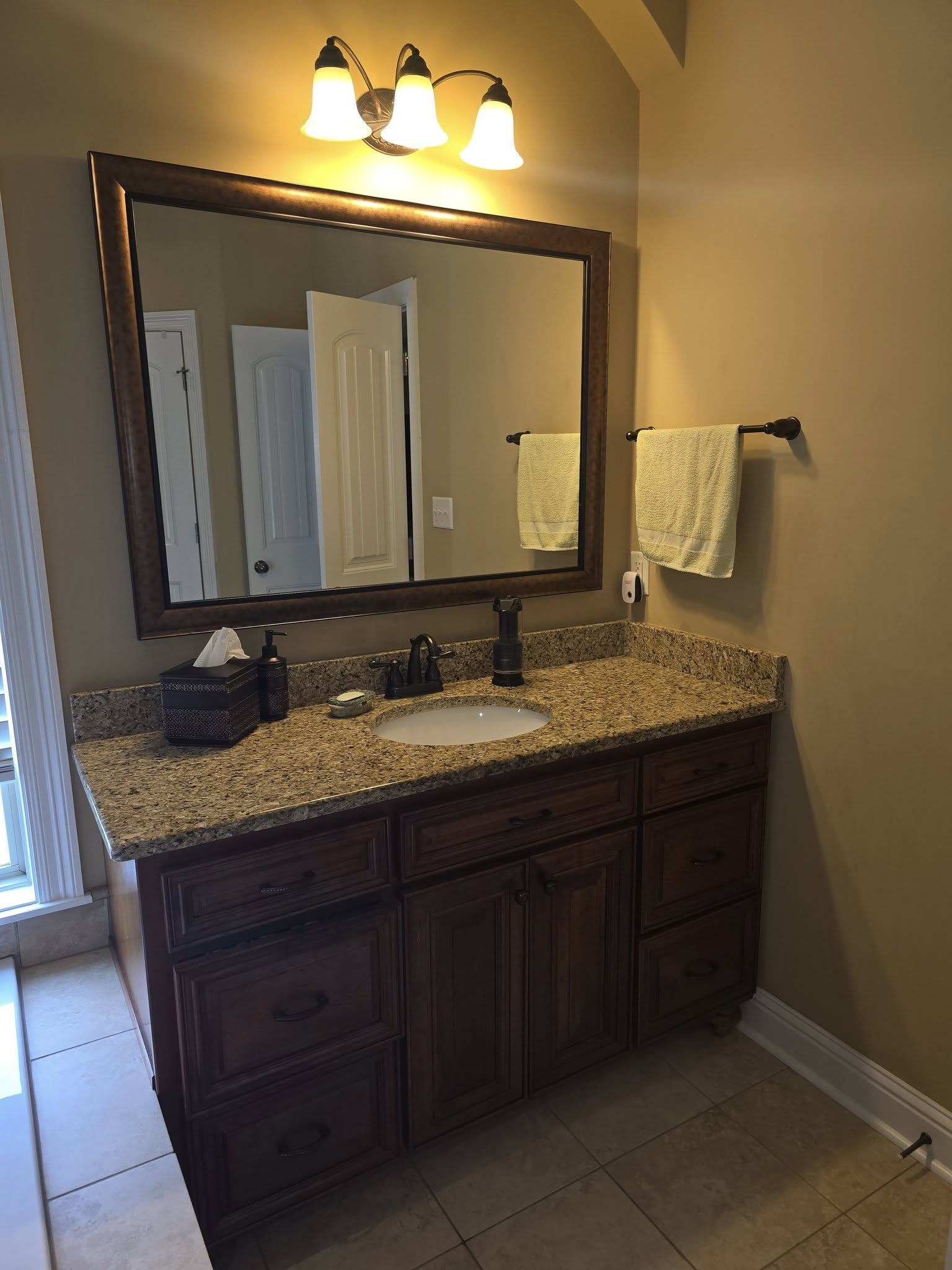 Bathroom vanity with brown cabinets, granite countertop, large mirror, and three-light fixture.