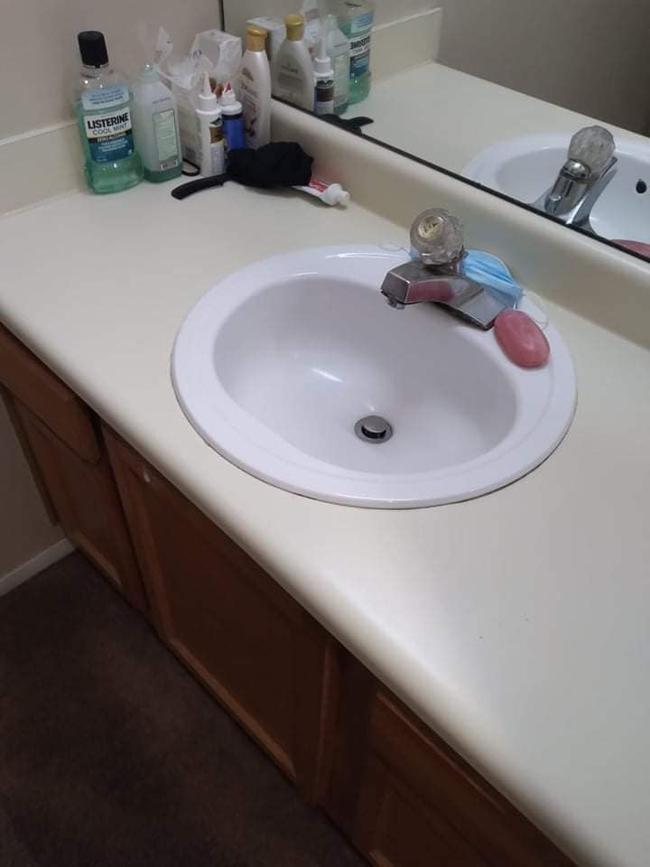 Bathroom sink with a white countertop and brown cabinets. Soaps, bottles, and a mirror are visible.