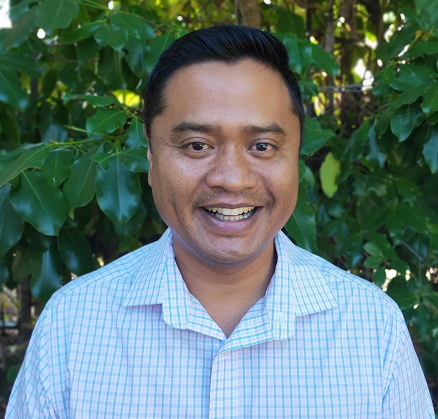 A man in a plaid shirt is smiling in front of a bush.