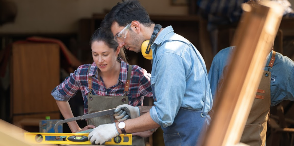 A man and a woman are working on a piece of wood in a workshop.