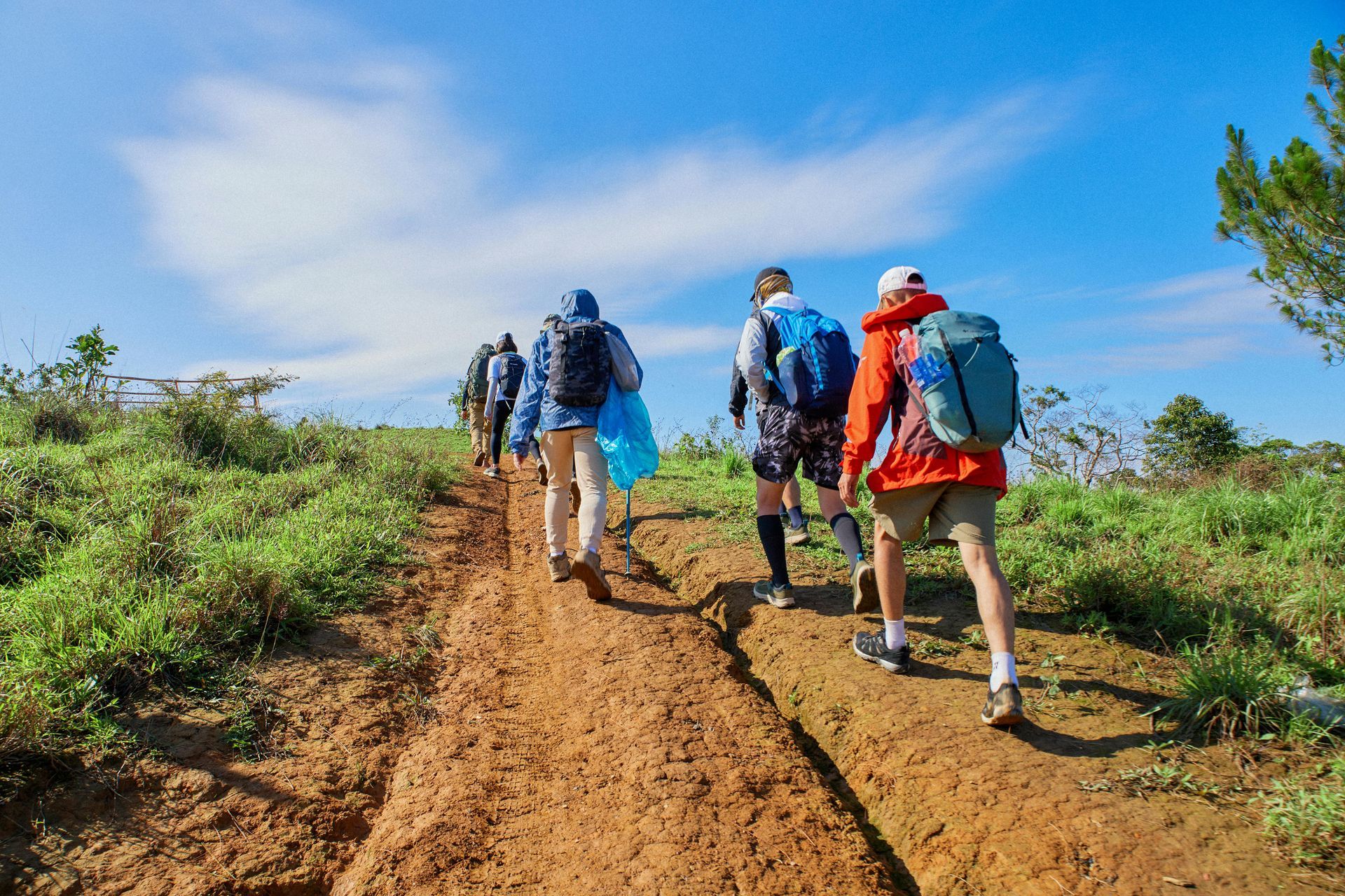 Hikers with backpacks walk uphill on a dirt path under a blue sky.