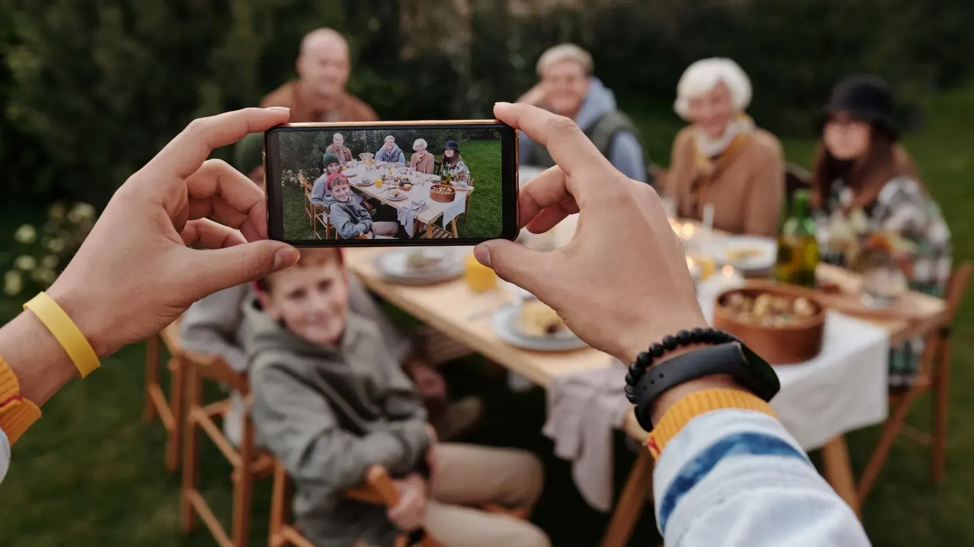 Hands holding a phone, taking a photo of a family gathered around a table outdoors.