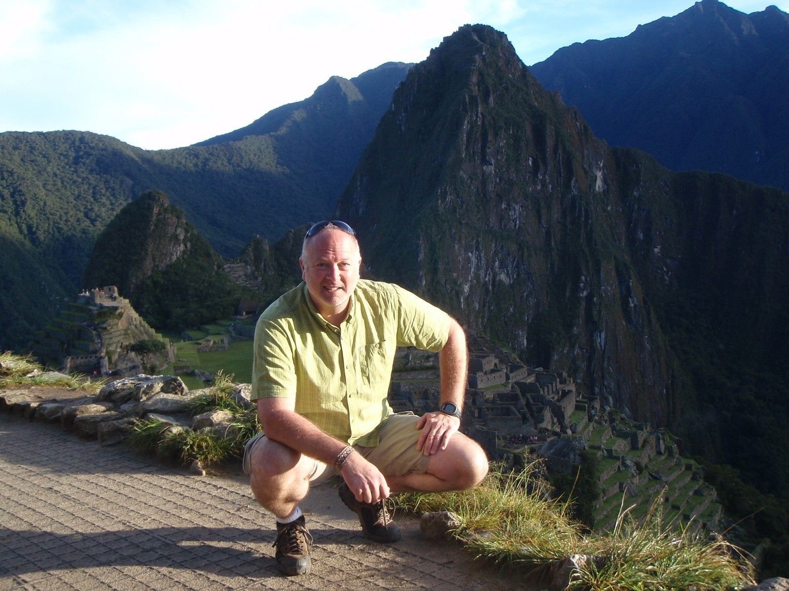 Man squatting with Machu Picchu ruins in the background. He wears light green shirt, beige shorts. Mountain setting.