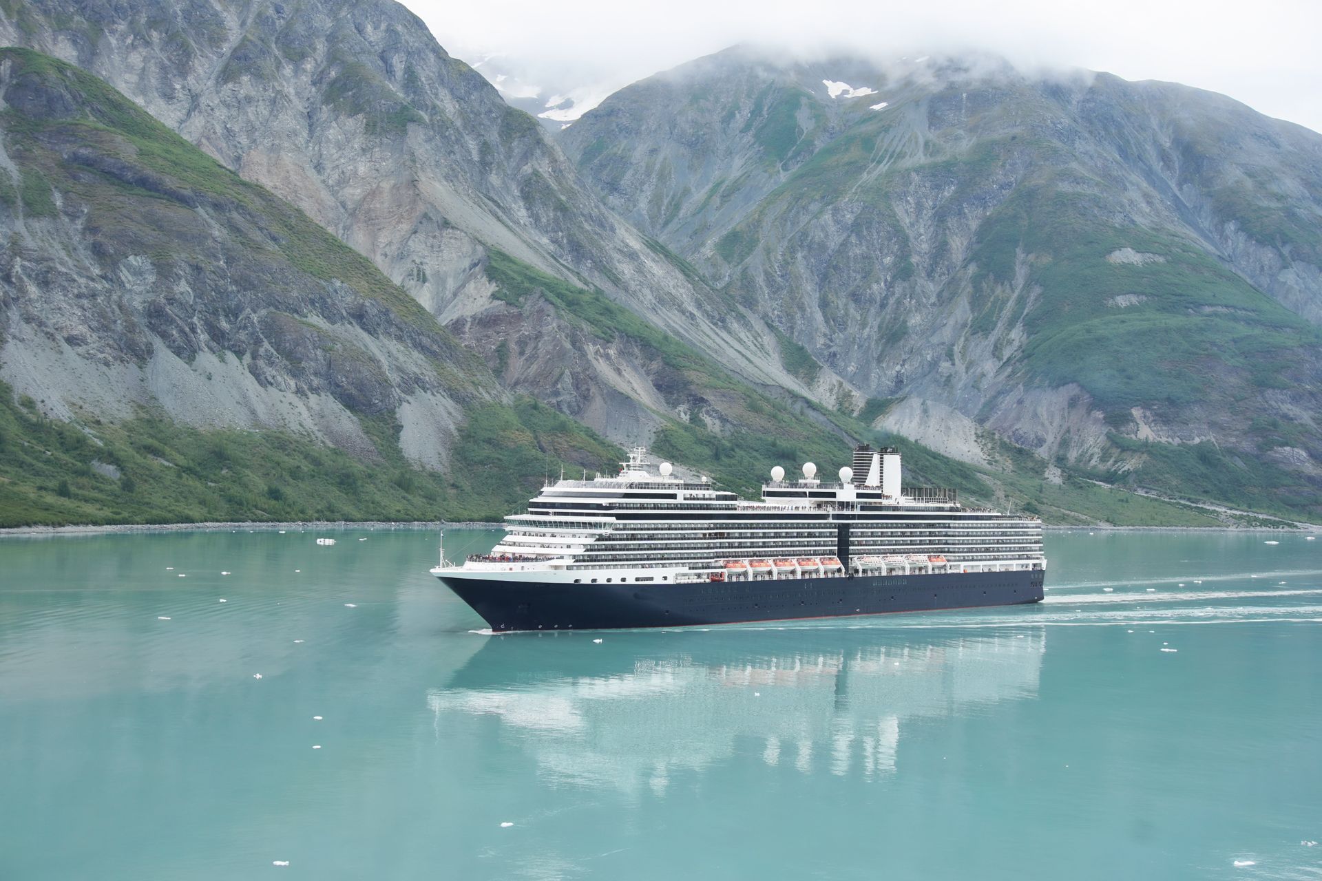 Cruise ship sails on turquoise water in front of mountains with a cloudy sky.