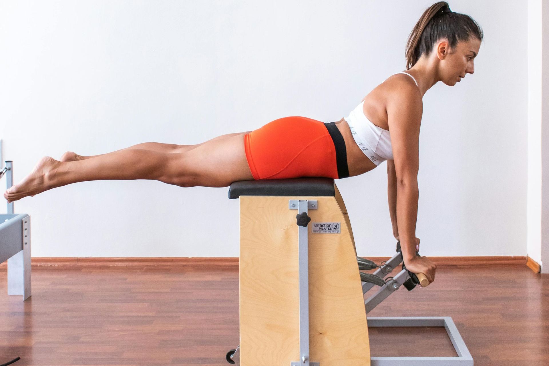 Woman doing Pilates exercise on a wooden chair, arms extended, legs lifted, wearing white top and orange shorts.