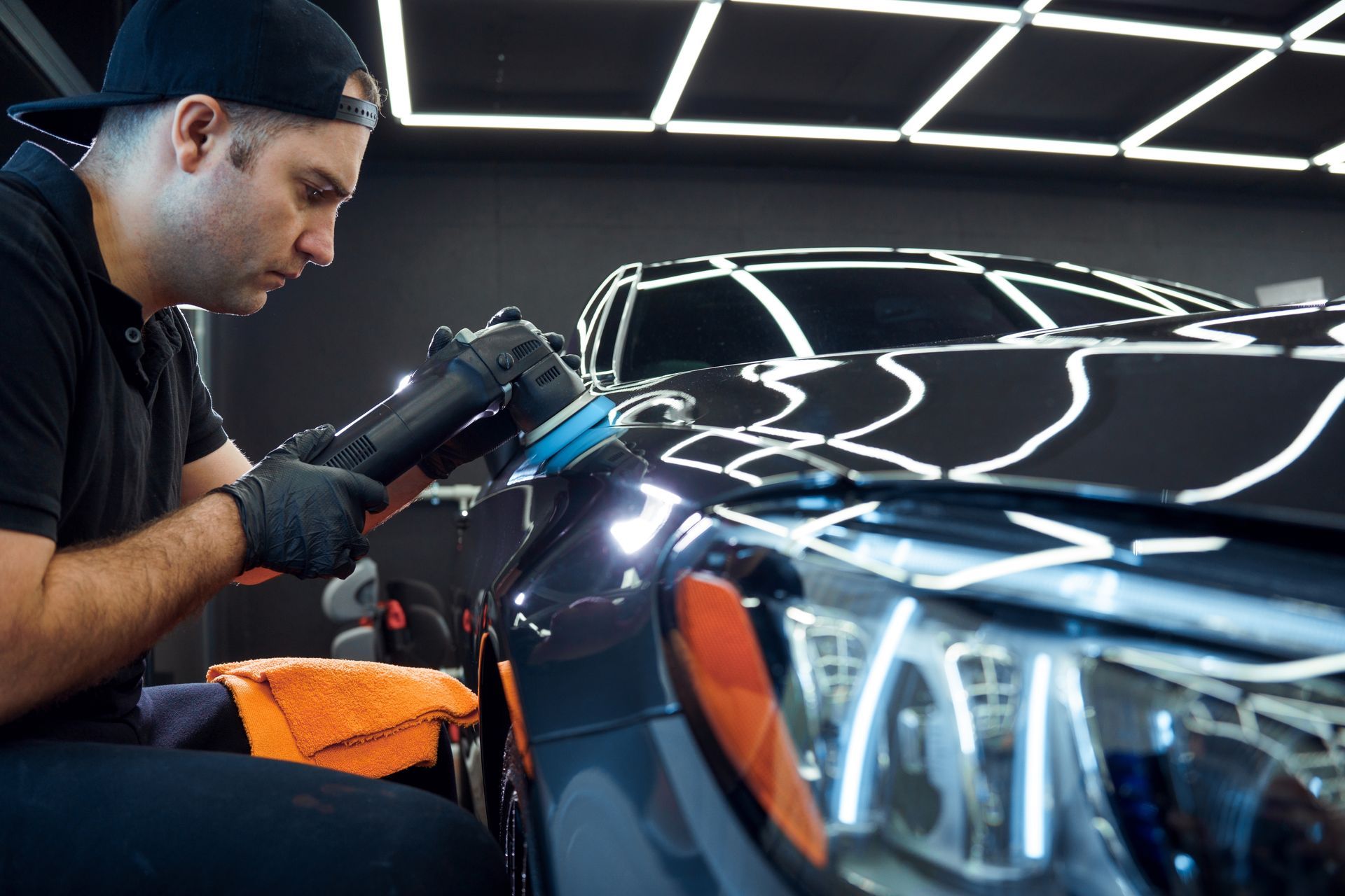 Man polishing a dark blue car, wearing a black hat and gloves, in a bright garage.