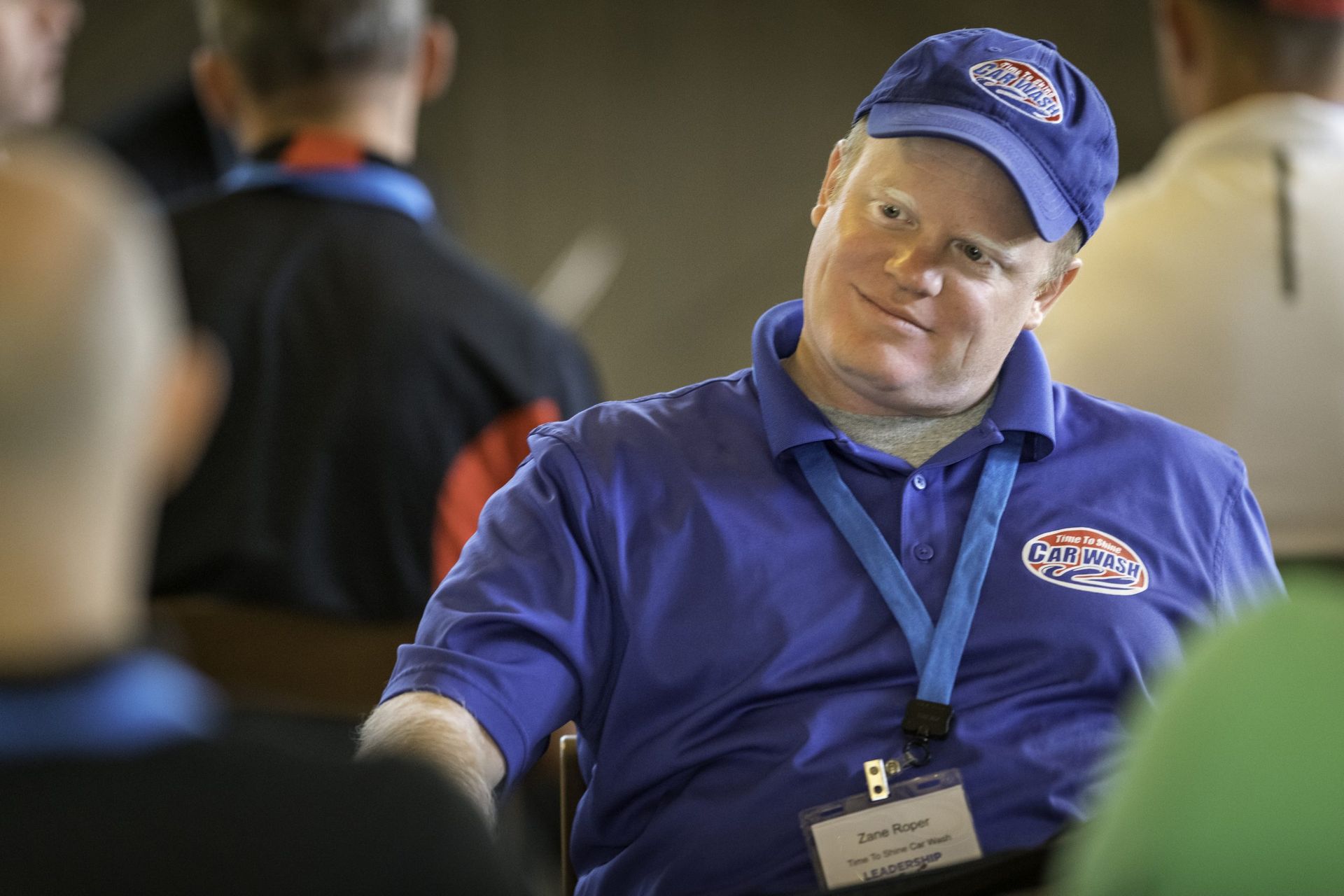 A man wearing a blue shirt and a blue hat is sitting at a table with other people.