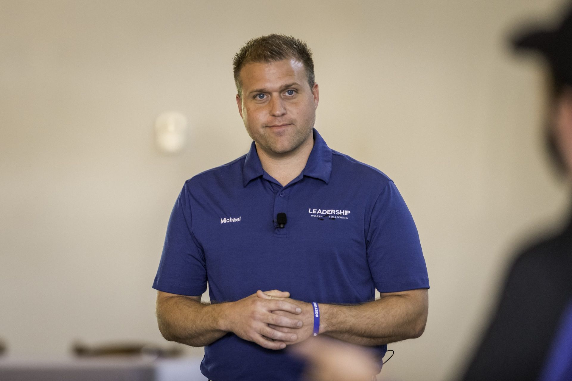A man in a blue shirt is standing in a room with his hands folded.
