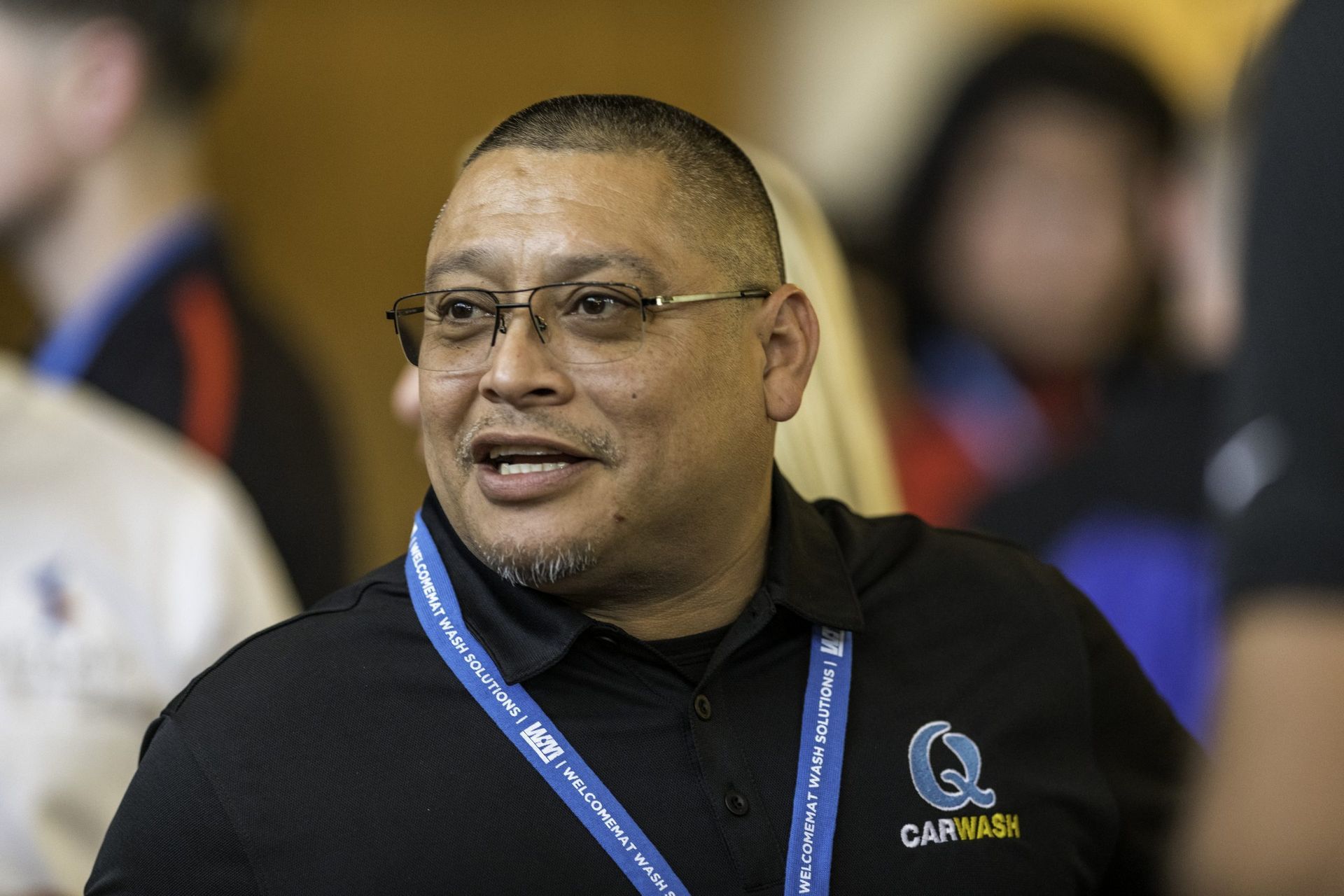 A man wearing glasses and a lanyard around his neck is smiling.
