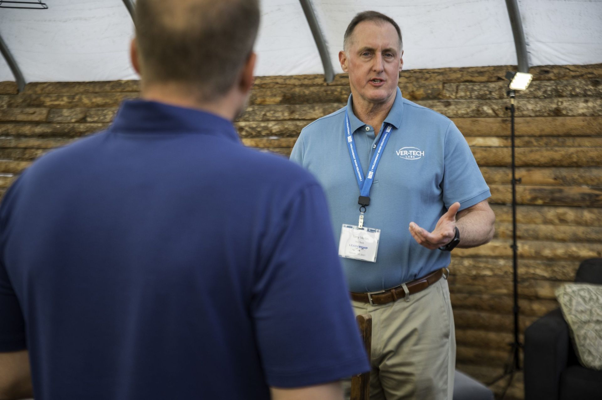 A man in a blue shirt is talking to another man in front of a wooden wall.