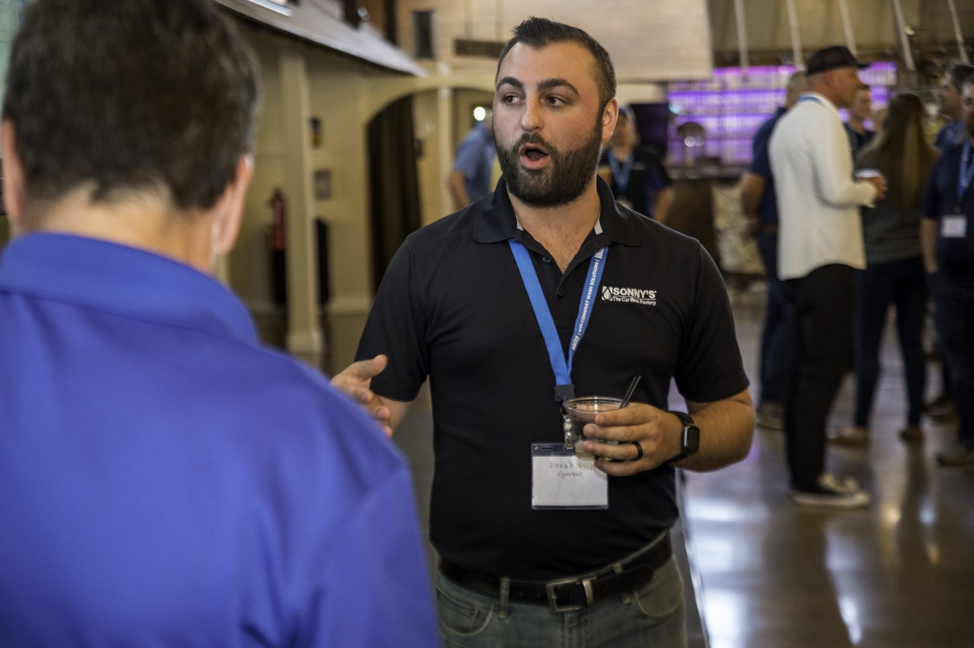 A man with a beard is talking to another man while holding a drink.