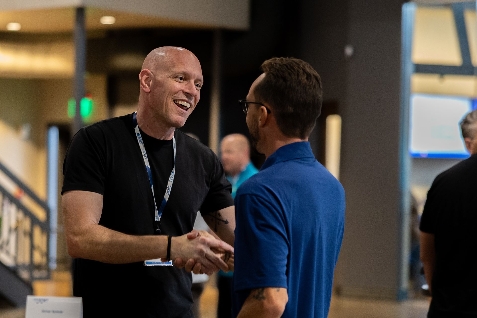 Two men shaking hands and laughing at Leadership Worth Following car wash convention . One man wears a black shirt, the other a blue polo.