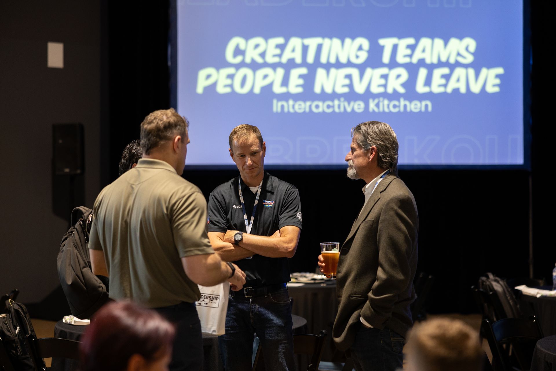 Three men conversing near a table with a screen behind them that says