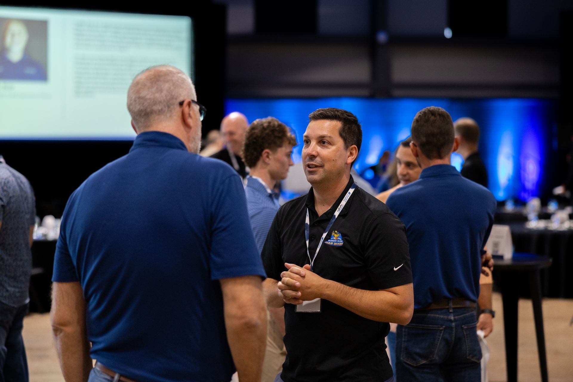 Two men in blue shirts converse at Leadership Worth Following car wash conference, others in background.