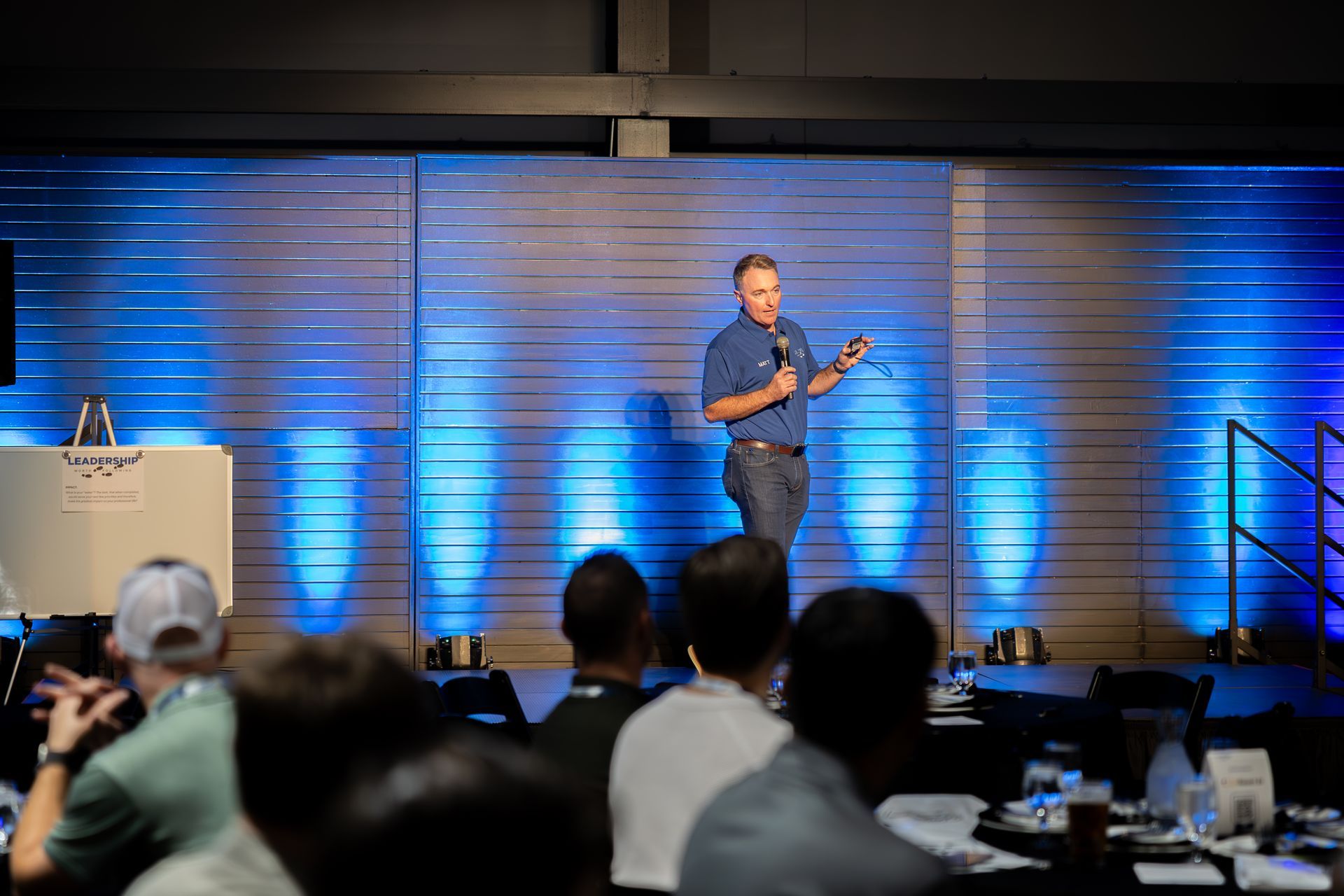 Man speaking at a leadership car wash conference. Stage lit with blue lights. Audience seated at tables.