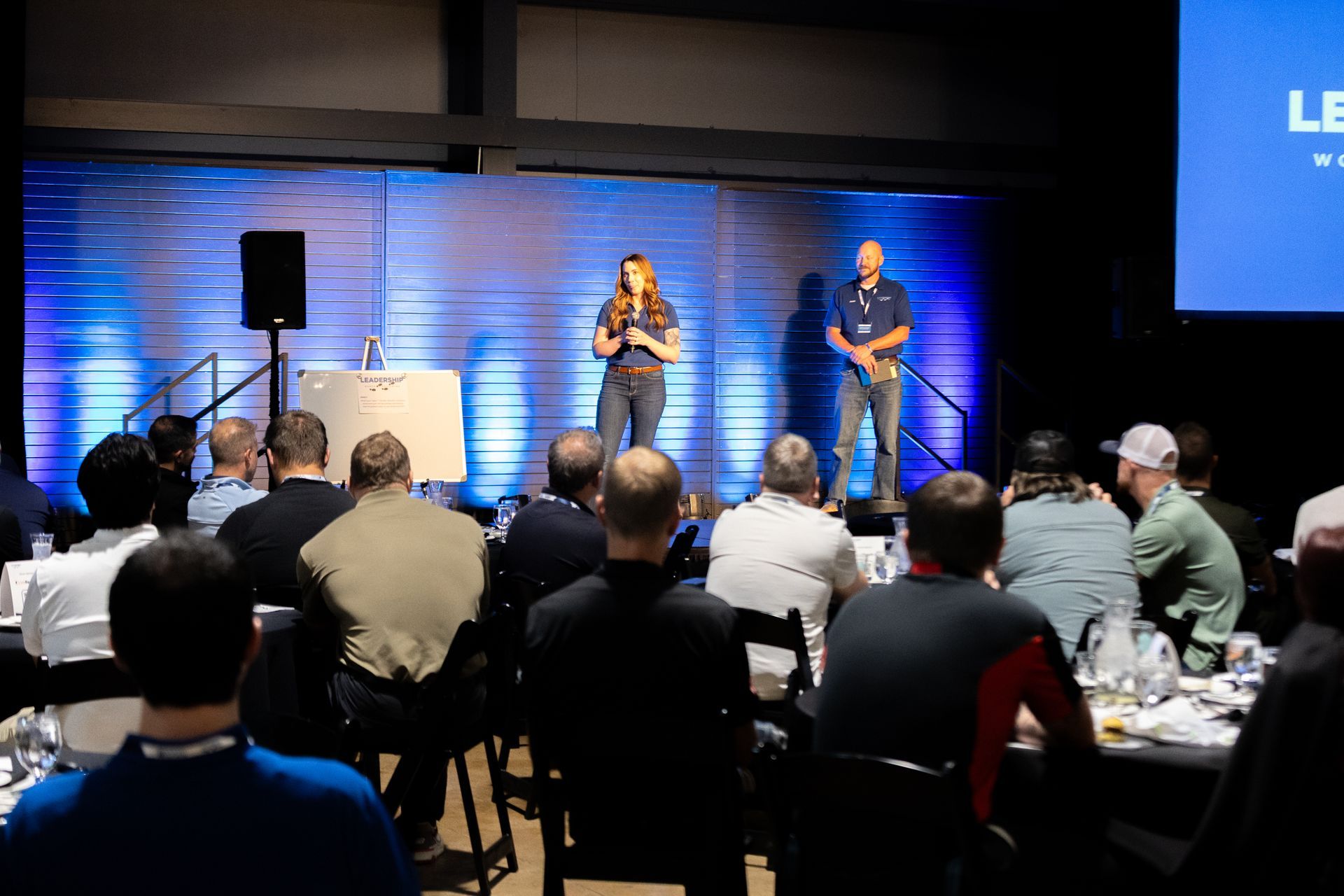 Two people speaking on a stage to an audience at Leadership Worth Following car wash conference. Blue background with a logo.