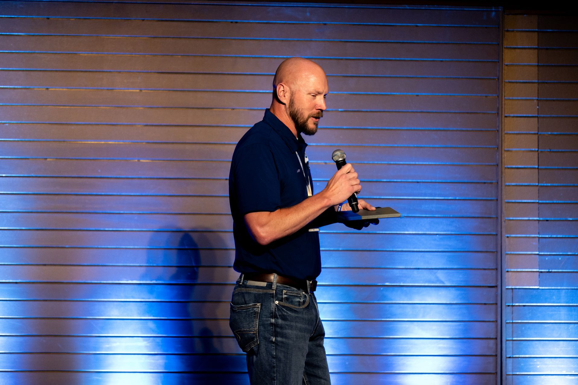 Man in dark blue shirt and jeans speaks at Leadership Worth Following car wash convention on a stage with blue and gray backdrop.