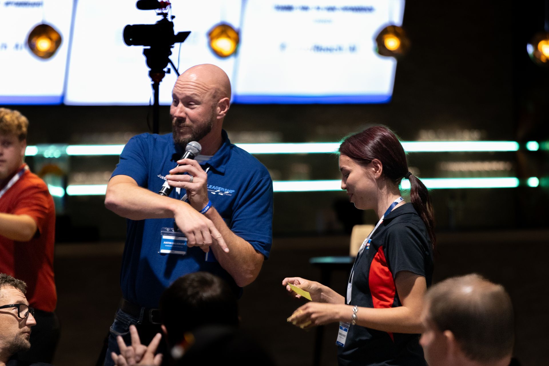 Man with microphone gestures, addressing a crowd at Leadership Worth Following car wash convention. A woman stands beside him.