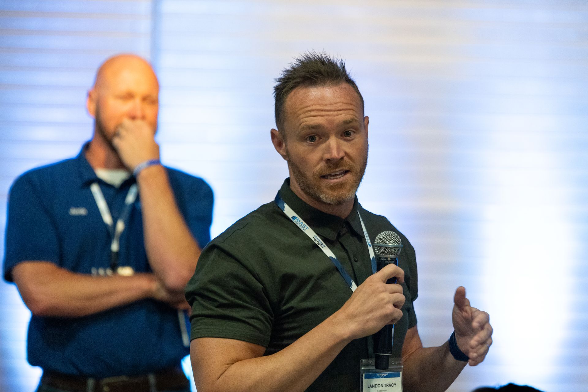 Man in dark green shirt speaking into a microphone at Leadership Worth Following car wash convention. Another man in blue shirt in the background, observing.